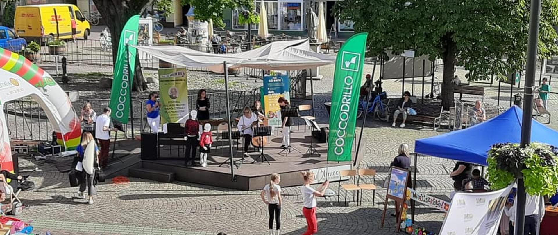 rynek w Lublińcu, zdjęcie robione z góry. Na środku pod drzewami Krzywa scena, na której nauczyciele szkoły muzycznej przygotowują się do koncertu, wokół kolorowe namioty, przy scenie kilkoro ludzi.