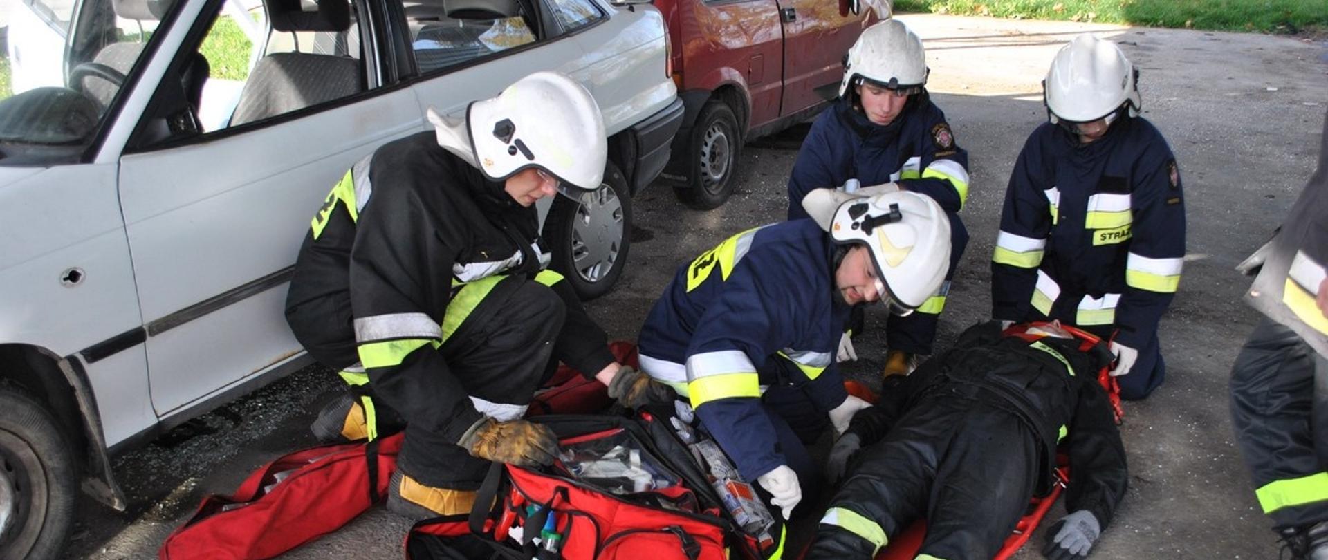 Na zdjęciu widać czterech ratowników szkolących się w udzielaniu pomocy poszkodowanemu. Osoba poszkodowana znajduję się na noszach typu deska, obok poszkodowanego znajduje się torba PSP-R1.