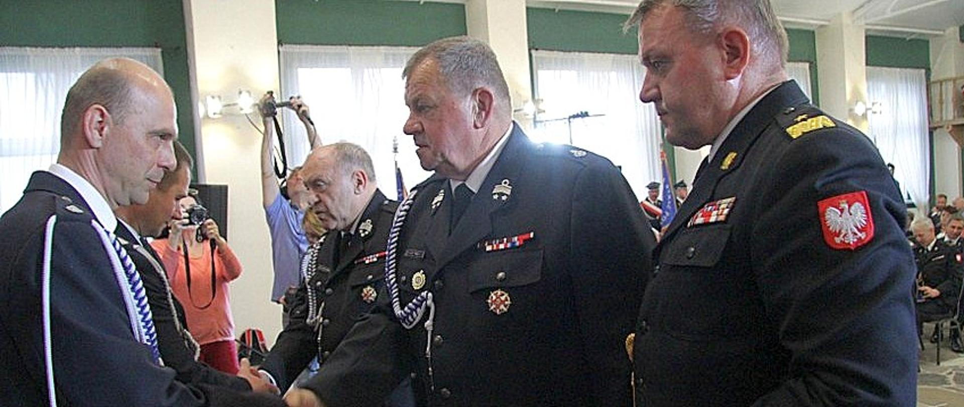 Zdjęcie zrobione wewnątrz pomieszczenia, w trakcie uroczystości. Druhowie ochotnicy oraz oficer Państwowej Straży Pożarnej w stopniu generała wręczają odznaczenia ustawionym w szeregu strażakom ochotnikom. 