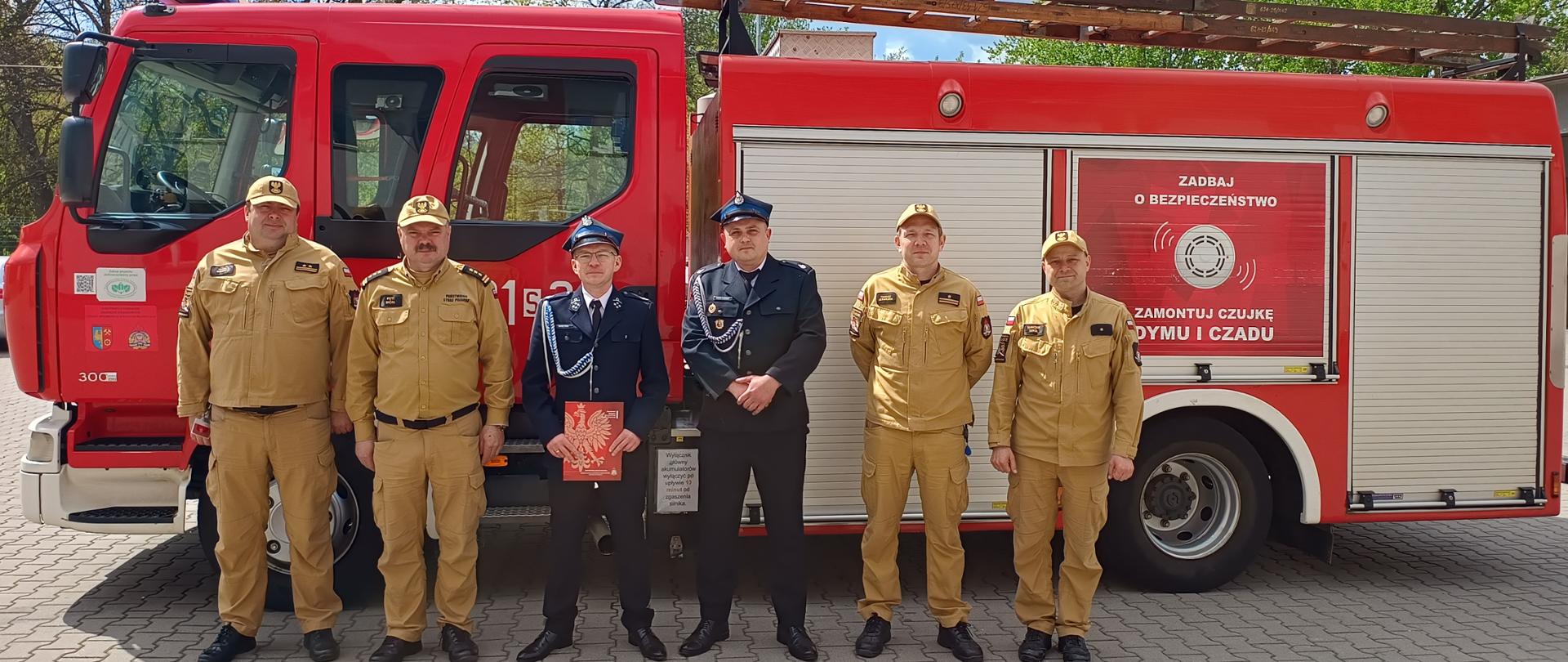 Na zdjęciu widać grupę sześciu strażaków pozujących do pamiątkowej fotografii na tle dużego, czerwonego wozu pożarniczego. Czterech mężczyzn ma na sobie piaskowe mundury służbowe z czapkami z daszkiem, natomiast dwaj pozostali ubrani są w ciemnogranatowe mundury wyjściowe z czapkami rogatywkami. Jeden ze strażaków w ciemnym mundurze trzyma przed sobą czerwoną teczkę z godłem Polski, co sugeruje, że zdjęcie dokumentuje ważne wydarzenie, takie jak przekazanie sprzętu, awans lub oficjalną wizytę.
Tło zdjęcia stanowi bok nowoczesnego samochodu gaśniczego, na którym zamontowano drabinę oraz roletę skrywającą sprzęt ratowniczy. Na bocznych drzwiach pojazdu widnieje hasło edukacyjne „Zadbaj o bezpieczeństwo. Zamontuj czujkę dymu i czadu”, promujące profilaktykę przeciwpożarową. Cała scena rozgrywa się na wybrukowanym placu przed jednostką straży pożarnej w słoneczny, wiosenny dzień, o czym świadczą wyraźne cienie i świeża zieleń drzew widoczna za pojazdem.
