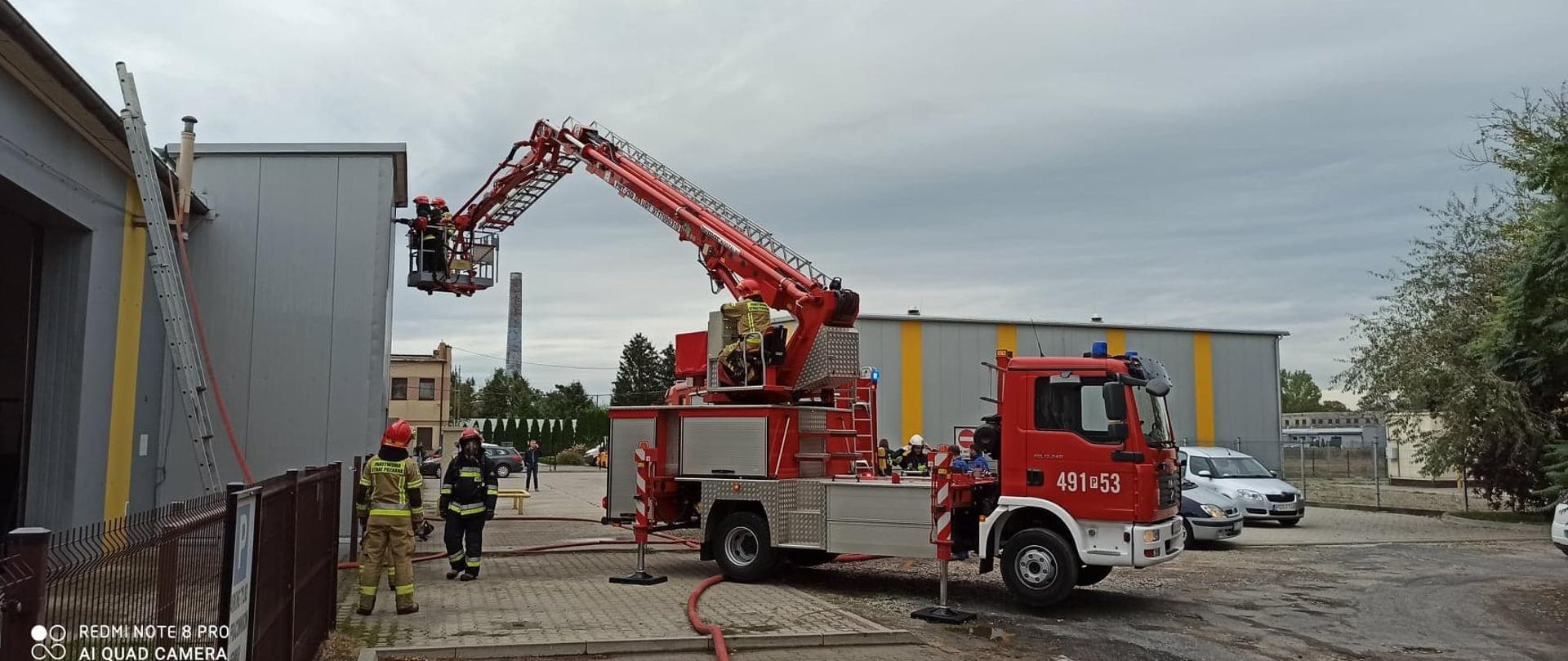 Przy hali produkcyjnej sprawiono podnośnik hydrauliczny. Dwóch strażaków w ubraniach bojowych, hełmach i sprzęcie ochrony dróg oddechowych uzyskują dostęp do powierzchni dachowej obiektu z kosza podnośnika celem weryfikacji zagrożenia kamerą termowizyjną. Dodatkowo w innej części hali sprawiono drabinę przystawną, po której poprowadzono linię gaśniczą na powierzchnie dachu. W pobliżu hali dwóch strażaków nadzoruje działania z podłoża. Za podnośnikiem hydraulicznym strażacy po zakończonej akcji gaśniczej zdejmują elementy ochrony osobistej. Działania toczą się na terenie zakładu produkcyjnego. W tle widoczne inne obiektu należące do firmy. 