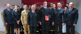 Świetlica komendy. Na tle ścianki promocyjnej do pamiątkowego zdjęcia pozuje awansowany strażak wraz z kierownictwem komendy oraz pozostałymi strażakami i pracownikami cywilnymi. W lewej ręce trzyma teczkę ze stosowną decyzją awansową. Strażacy mają na sobie mundury wyjściowe oraz służbowe w kolorze piaskowym i koszarowe.