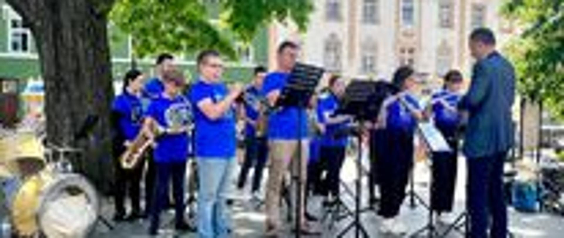 Zdjęcie przedstawia grupę muzyków grających na instrumentach oraz dyrygenta, na podeście, w tle budynek historyczny