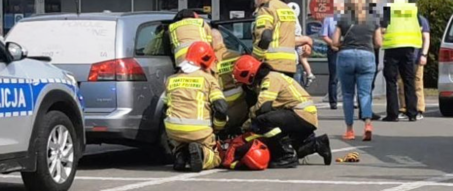 Strażacy udzielający pierwszej pomocy kobiecie znajdującej się w samochodzie osobowym, na pierwszym planie przód radiowozu policyjnego, w oddali policjant i osoby cywilne.