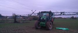 Zdjęcie przedstawia pole, na którym w części centralnej znajduje się ciągnik z opryskiwaczem. Po lewej stronie znajduje się złamany słup energetyczny. Po prawej stronie ciągnika na ziemi leży osoba poszkodowana, która poniosła śmierć w skutek porażenia prądem elektrycznym. 