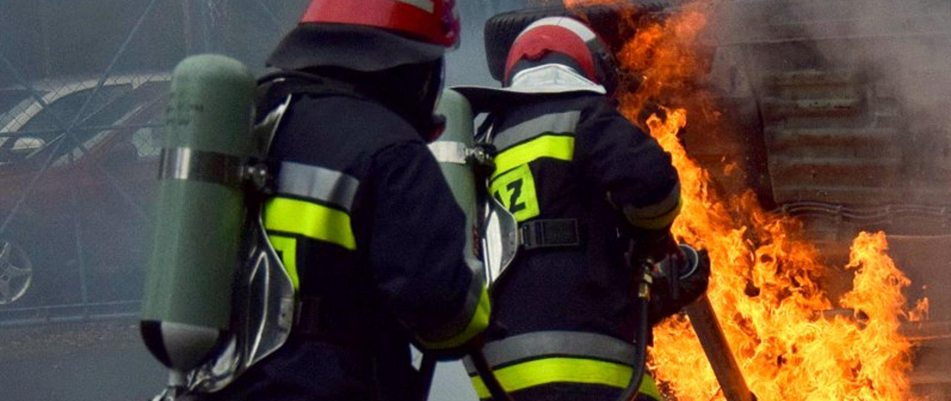 Dwóch strażaków ubranych w ubrania specjalne (nomexy) w hełmach i aparatach powietrznych gasi podwozie płonącego auta. Samochód leży na boku