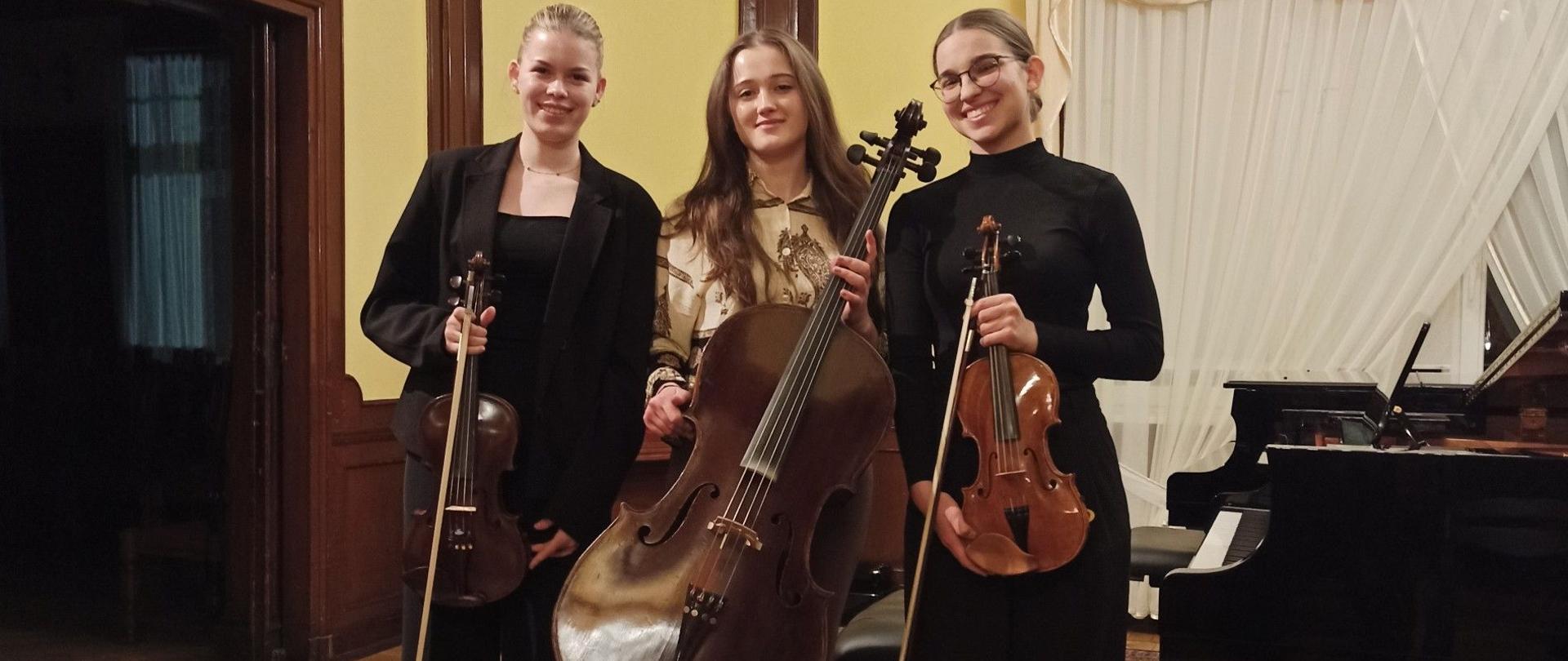 
Treść wiadomości Gemini
Zdjęcie kolorowe. Trzy kobiety stojące obok siebie w eleganckim wnętrzu z instrumentami smyczkowymi. Kobieta po lewej w czarnym garniturze trzyma skrzypce i smyczek. Kobieta w środku w jasnej, wzorzystej koszuli trzyma wiolonczelę. Kobieta po prawej w okularach i czarnym golfie trzyma skrzypce i smyczek. W tle drewniane panele ścienne, fragment fortepianu, jasne zasłony oraz wzorzysty dywan na podłodze.