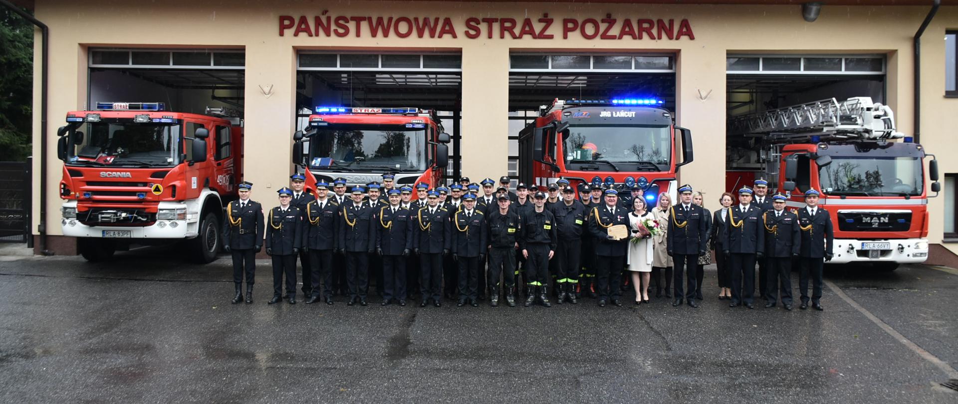Zdjęcie przedstawia plac zewnętrzny Komendy Powiatowej. Na pierwszym planie przed budynkiem Komendy stoją pracownicy oraz funkcjonariusze Państwowej Straży Pożarnej w Łańcucie w pamiątkowym zdjęciu. W tle garaże komendy oraz pojazdy Komendy.