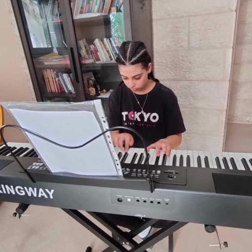 Nastolatek siedzi przy przenośnym pianinie ustawionym na tle szafy z książkami i gra z nut zapisanych na kartce położonej na pulpicie