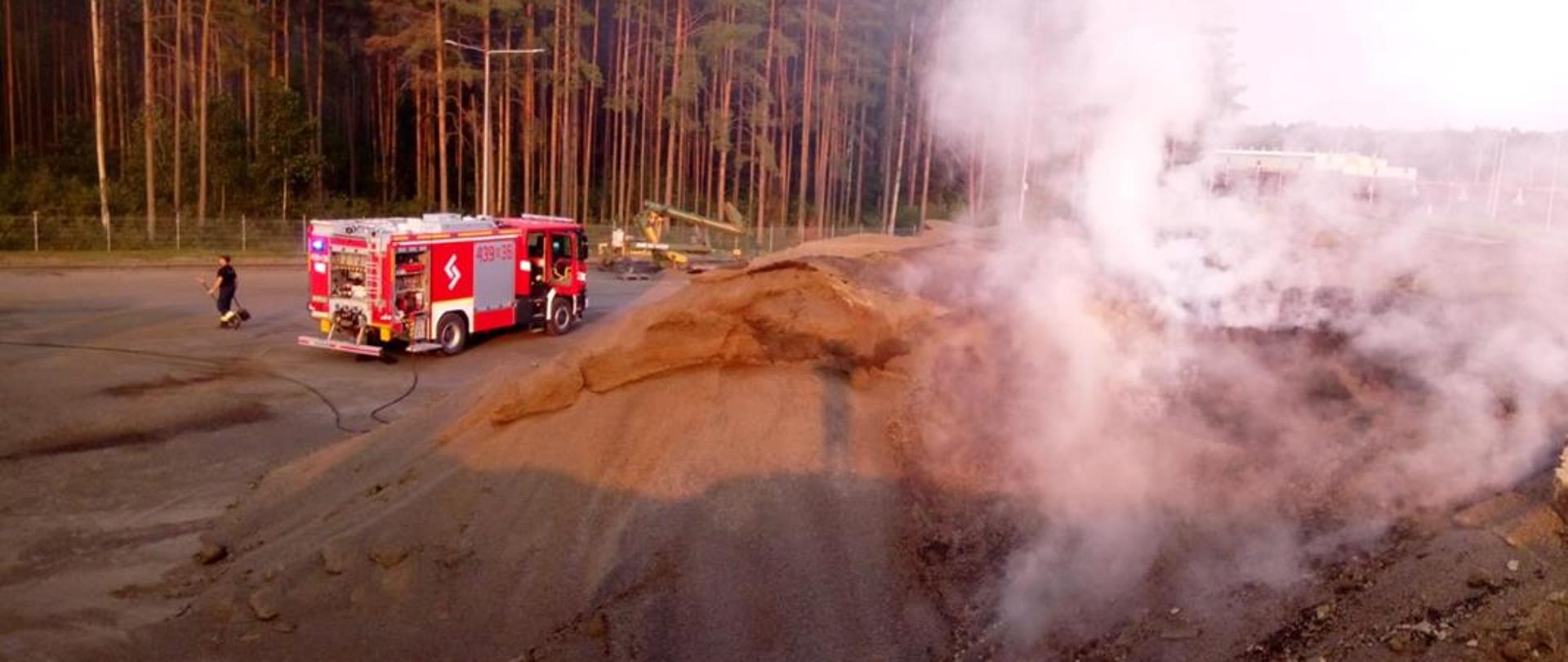 Na pierwszym planie hałda palącego się pelletu z widocznym zadymieniem. Na planie drugim samochód ratowniczo-gaśniczy z rozwiniętą linią gaśniczą.