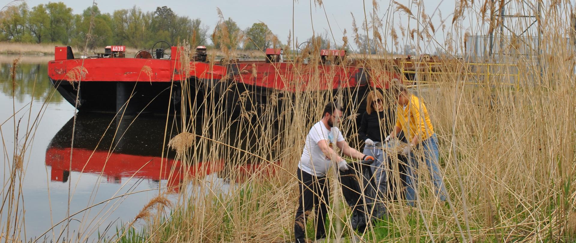 Sprzątanie brzegu rzeki Odra. Troje ludzi zbiera śmieci w tle na nabrzeżu stoi przycumowana czarno czerwona barka.