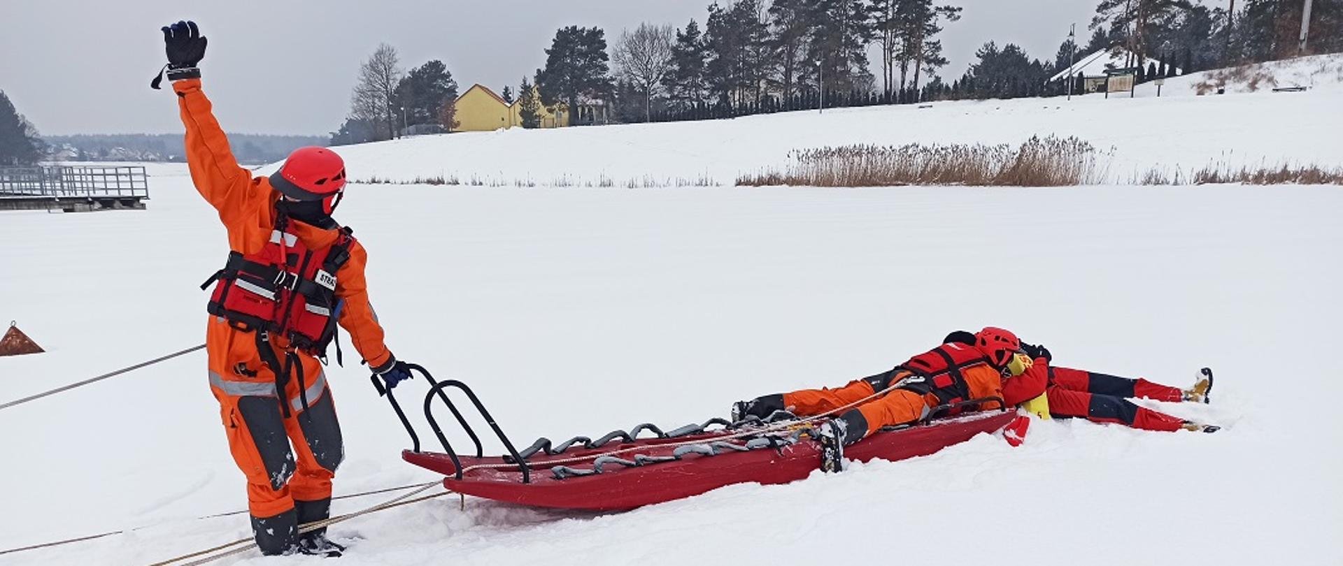 Na zdjęciu widać trzech ratowników na zamarzniętym akwenie wodnym. Zamarznięty akwen pokryty jest warstwą śniegu. Na pierwszym planie zdjęcia widać jak jeden z ratowników trzyma jedną ręką uchwyt sani lodowych natomiast drugą ma uniesioną w górę. Drugi z ratowników leży na saniach lodowych i podejmuje tonącego z przerębla. Wszyscy ratownicy ubrani są w pomarańczowe kombinezony i kaski. W tle widać linię brzegową porośniętą trzciną. Widoczne są również w oddali pojedyncze budynki mieszkalne oraz drzewa.