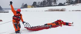 Na zdjęciu widać trzech ratowników na zamarzniętym akwenie wodnym. Zamarznięty akwen pokryty jest warstwą śniegu. Na pierwszym planie zdjęcia widać jak jeden z ratowników trzyma jedną ręką uchwyt sani lodowych natomiast drugą ma uniesioną w górę. Drugi z ratowników leży na saniach lodowych i podejmuje tonącego z przerębla. Wszyscy ratownicy ubrani są w pomarańczowe kombinezony i kaski. W tle widać linię brzegową porośniętą trzciną. Widoczne są również w oddali pojedyncze budynki mieszkalne oraz drzewa.