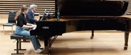 Nastolatka gra na fortepianie, za nią przy drugim fortepianie siedzi mężczyzna.