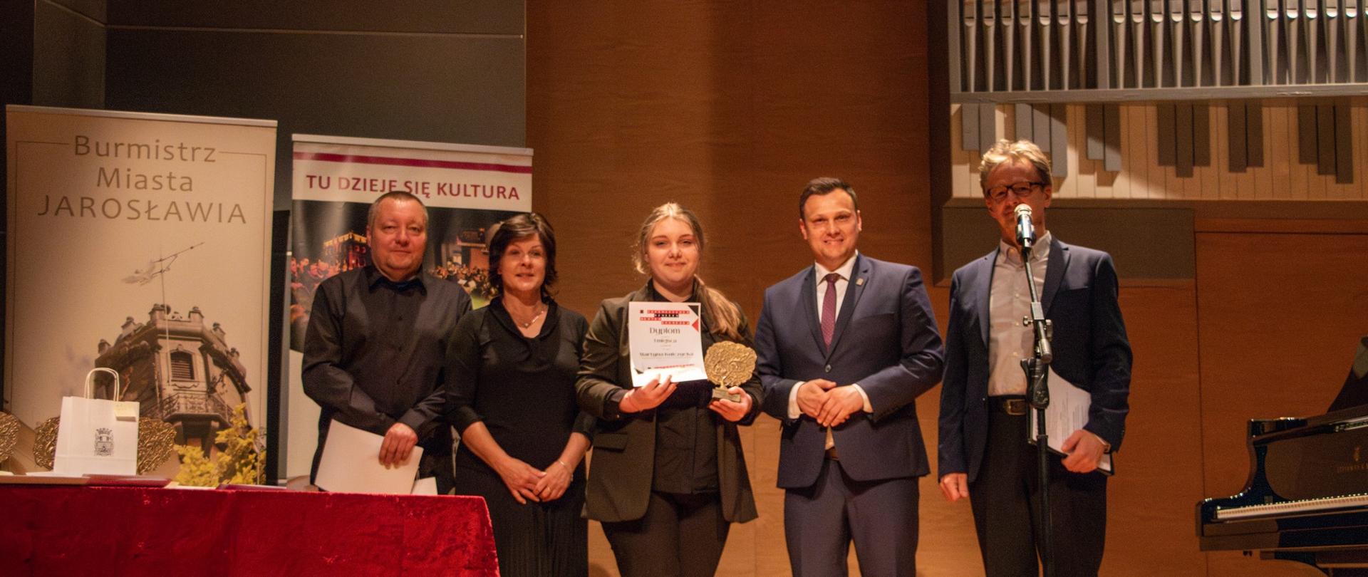 Alternatywny opis zdjęcia:
Na drewnianej scenie stoi pięć osób za stołem przykrytym czerwoną tkaniną. Od lewej stoi mężczyzna w czarnej koszuli, za nim częściowo widoczny jest baner z napisem "Burmistrz Miasta Jarosławia". Następnie stoi kobieta w czarnej sukience. Obok niej znajduje się młoda kobieta z długimi, jasnymi włosami, trzymająca w rękach dyplom i złoty medal. Dalej stoi mężczyzna w ciemnym garniturze i bordowym krawacie, a na końcu po prawej stronie stoi mężczyzna w szarej marynarce i ciemnych spodniach, trzymający mikrofon na statywie i kartkę papieru.
W tle widoczna jest ściana z drewnianą boazerią, a nad nią fragmenty organów piszczałkowych oraz baner informujący o "Ogólnopolskim Konkursie Muzyki Polskiej" w Jarosławiu w 2025 roku. Po prawej stronie kadru widoczny jest fragment czarnego fortepianu. Oświetlenie sceniczne skupia się na grupie osób stojących na scenie.