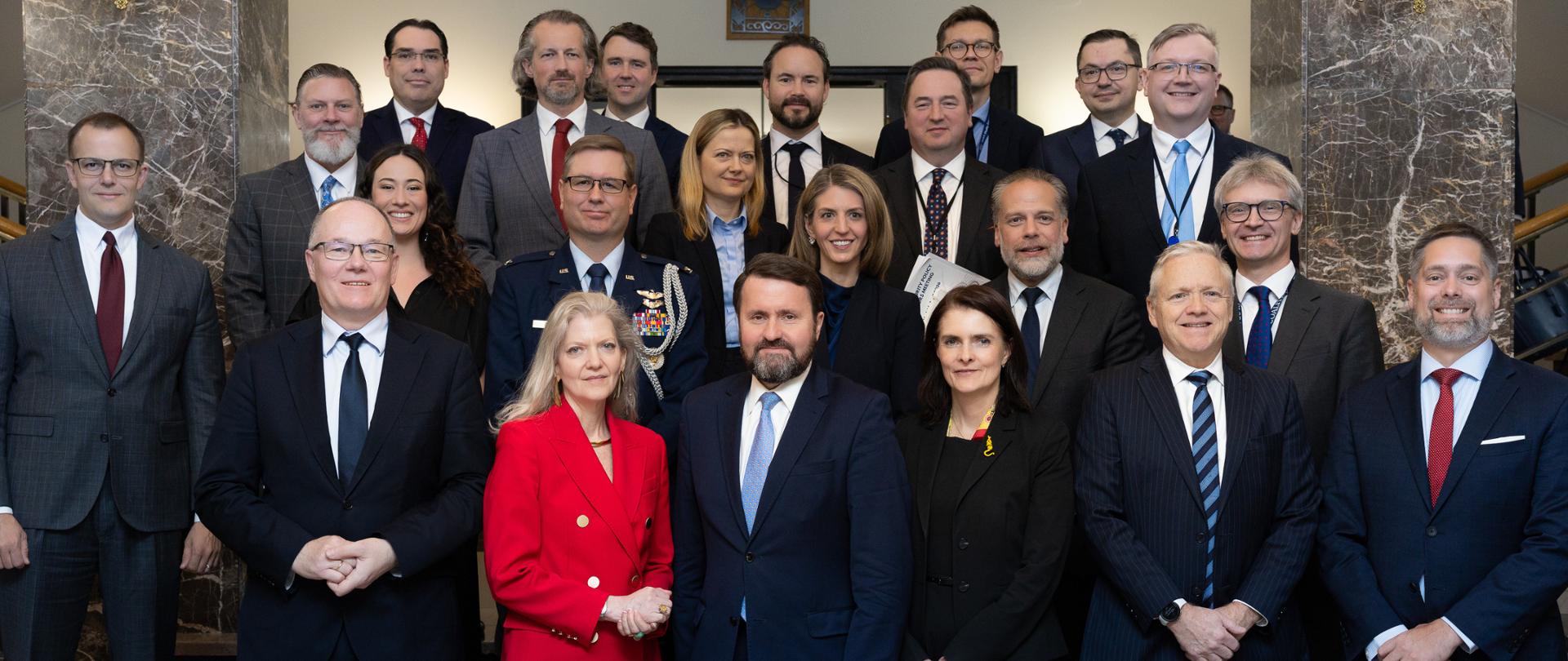 Grupa osób pozuje do wspólnego zdjęcia w reprezentacyjnym wnętrzu; uczestnicy ubrani są głównie w formalne stroje – garnitury i garsonki, w tym jedna kobieta w czerwonym kostiumie - podsekretarz stanu USA ds. politycznych Allison Hooker. Obok niej podsekretarz stanu w Ministerstwie Spraw Zagranicznych Robert Kupiecki.