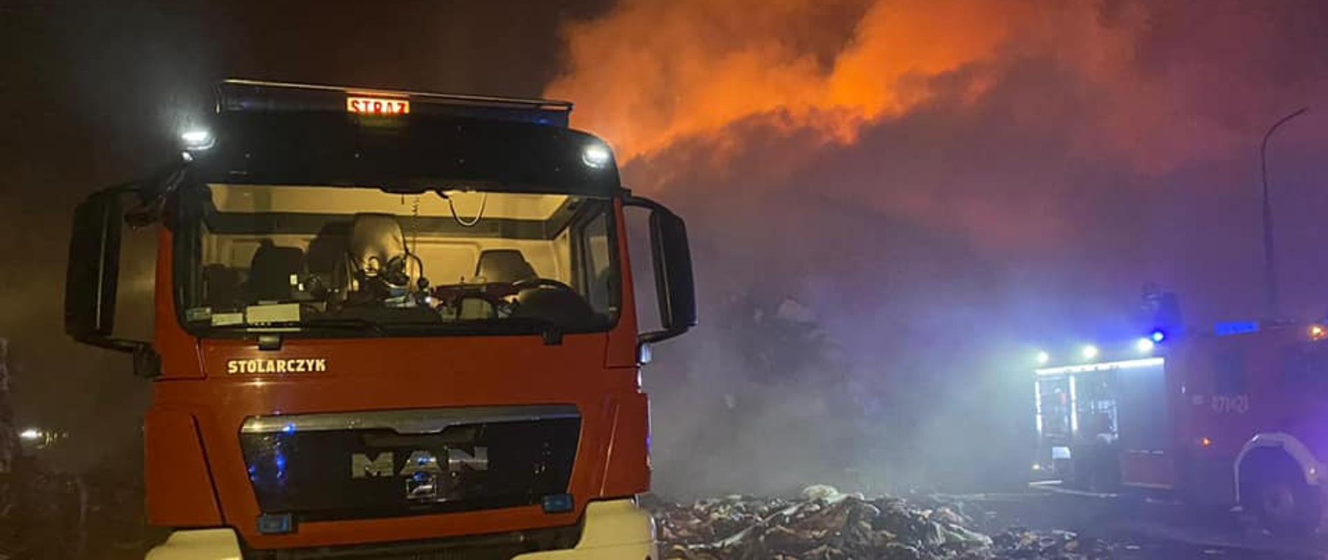Zdjęcie przedstawia ciężki samochód gaśniczy, biegnące linie wężowe oraz pożar pryzmy materiałów tekstylnych. W tle widoczny również średni samochód gaśniczy.