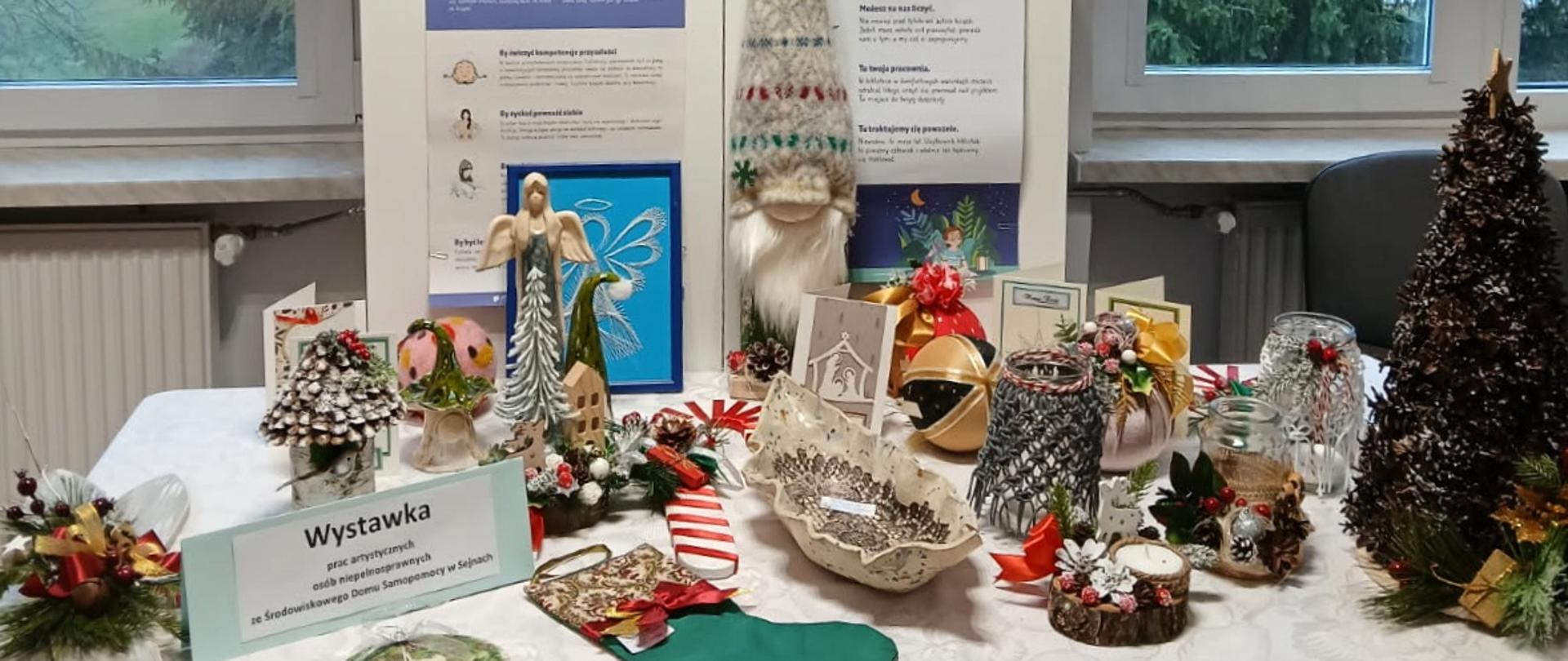 Stolik z wystawą świątecznych dekoracji i rękodzieła, w tym figurek, stroików i ozdób, prezentowany na tle okien i plakatu informacyjnego w pomieszczeniu biblioteki. Na pierwszym planie widoczna kartka z napisem „Wystawka prac artystycznych osób niepełnosprawnych ze Środowiskowego Domu Samopomocy w Sejnach”.