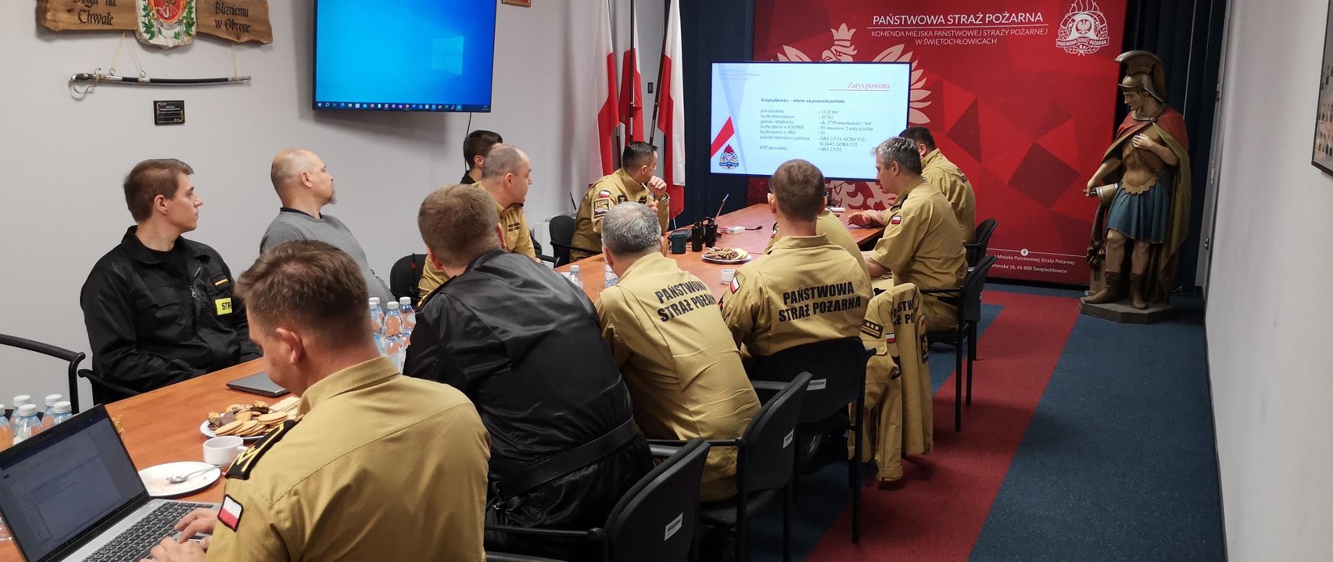 W sali w komendzie, po obudwu stronach siedzą w mundurach służbowych piaskowych i czarnych strażacy. Strażak na pierwszym planie pisze na laptopie. W tle ekran z prezentacją. Po prawej stronie rzeźba świętego Floriana. Po lewej flagi państwowe.
