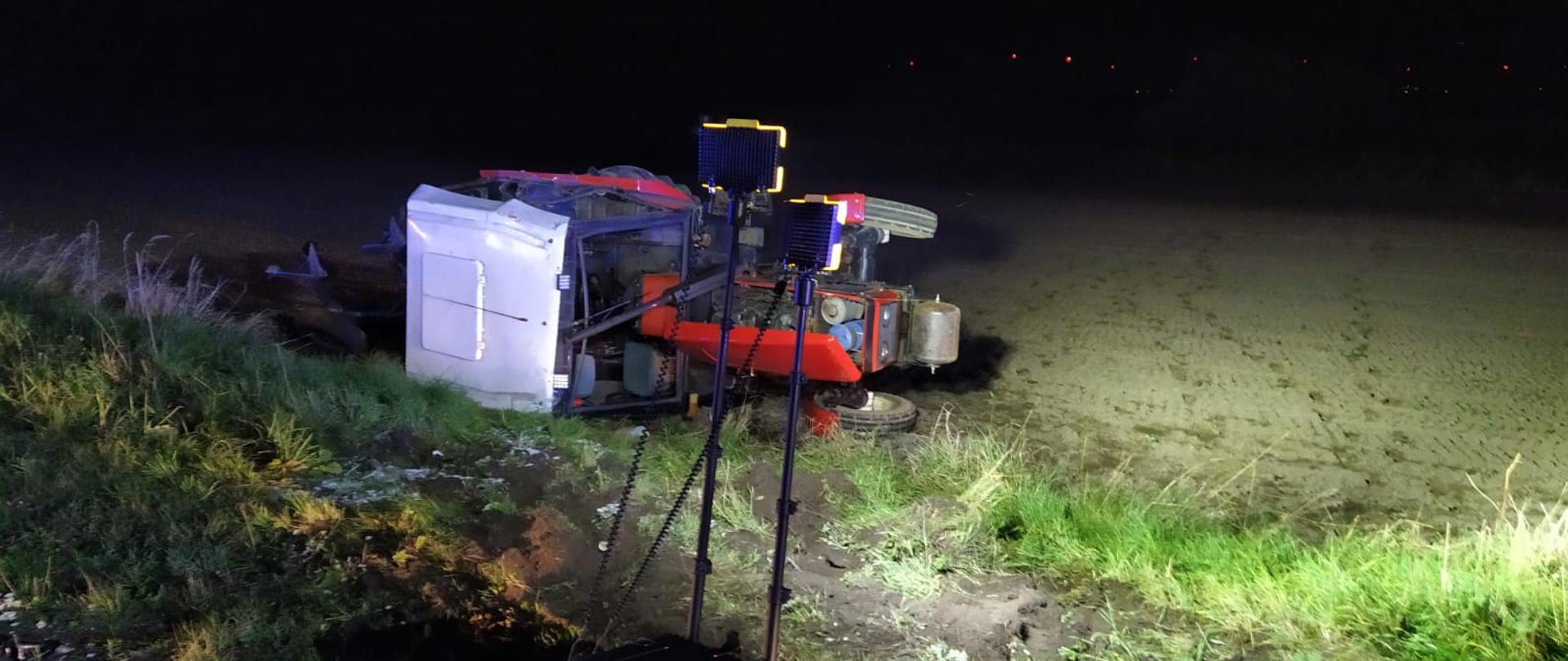 Obraz przedstawia leżący na prawym boku traktor w porze wieczornej. Traktor leży poza jezdnią na polu. Teren oświetla lampa.
