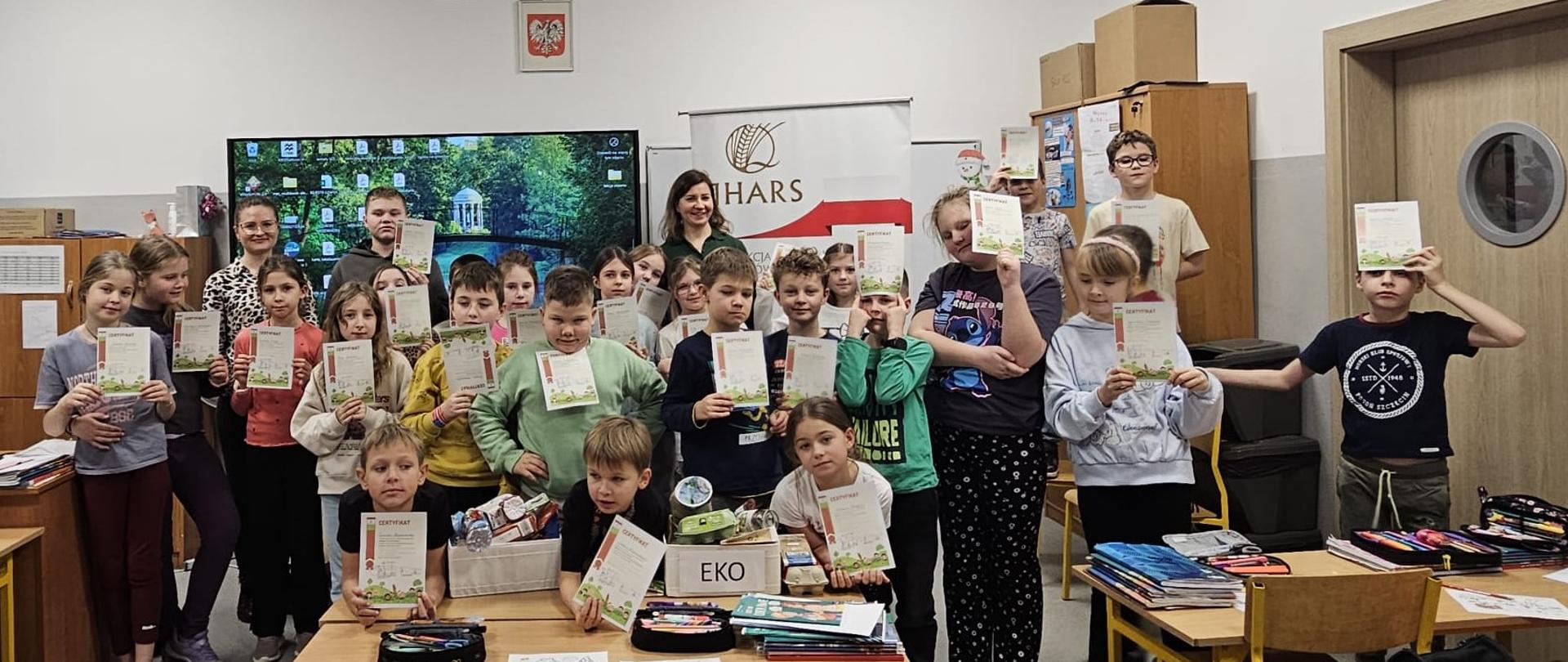 Inspektorka IJHARS stojąca z grupą dzieci trzymających dyplomy; wspólne zdjęcie grupowe na tle tablicy multimedialnej i baneru IJHARS.