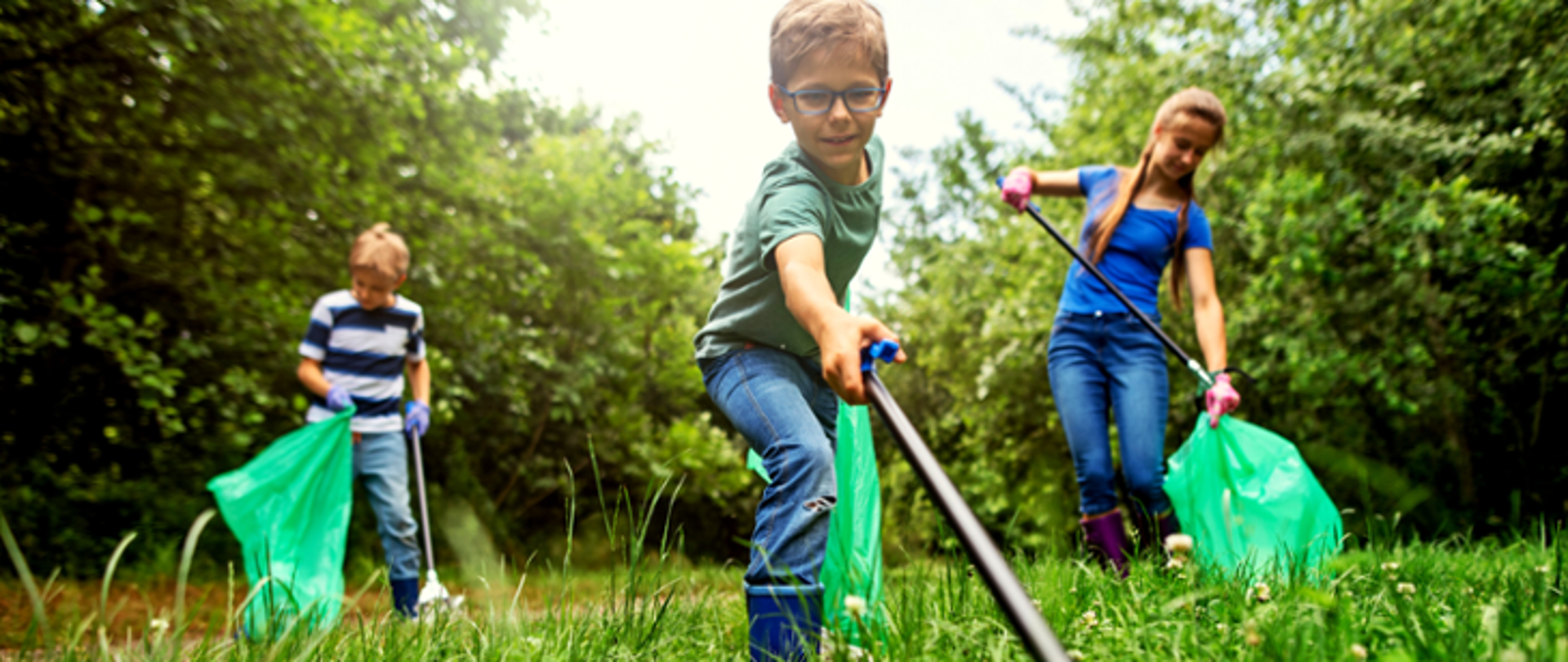 Zielona łąka, w tle krzaki. Troje dzieci dwóch chłopców i dziewczyna zbierają śmieci do worków.