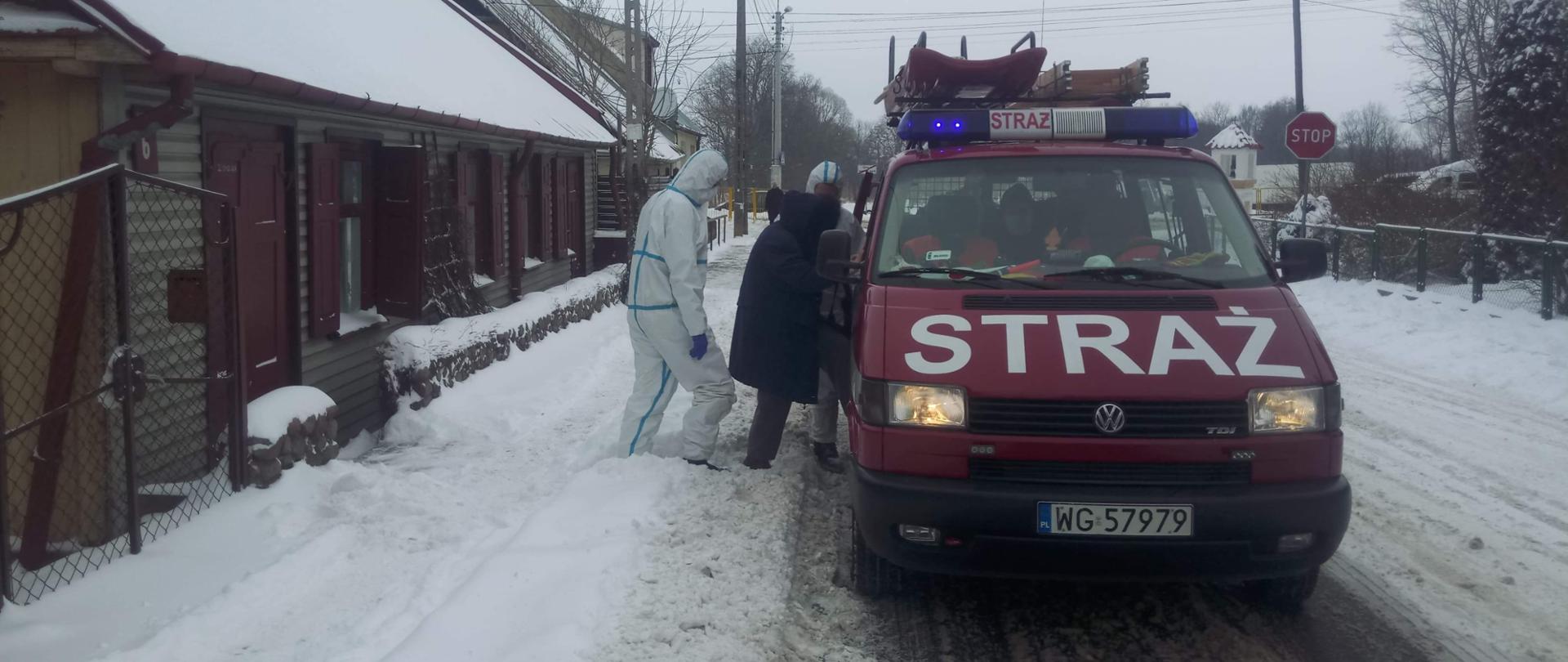 samochód strażacki przed którym stoi człowiek ubrany w biały kombinezon ochronny. poza zimowa. przed budynkiem stoi mężczyzna w maseczce.