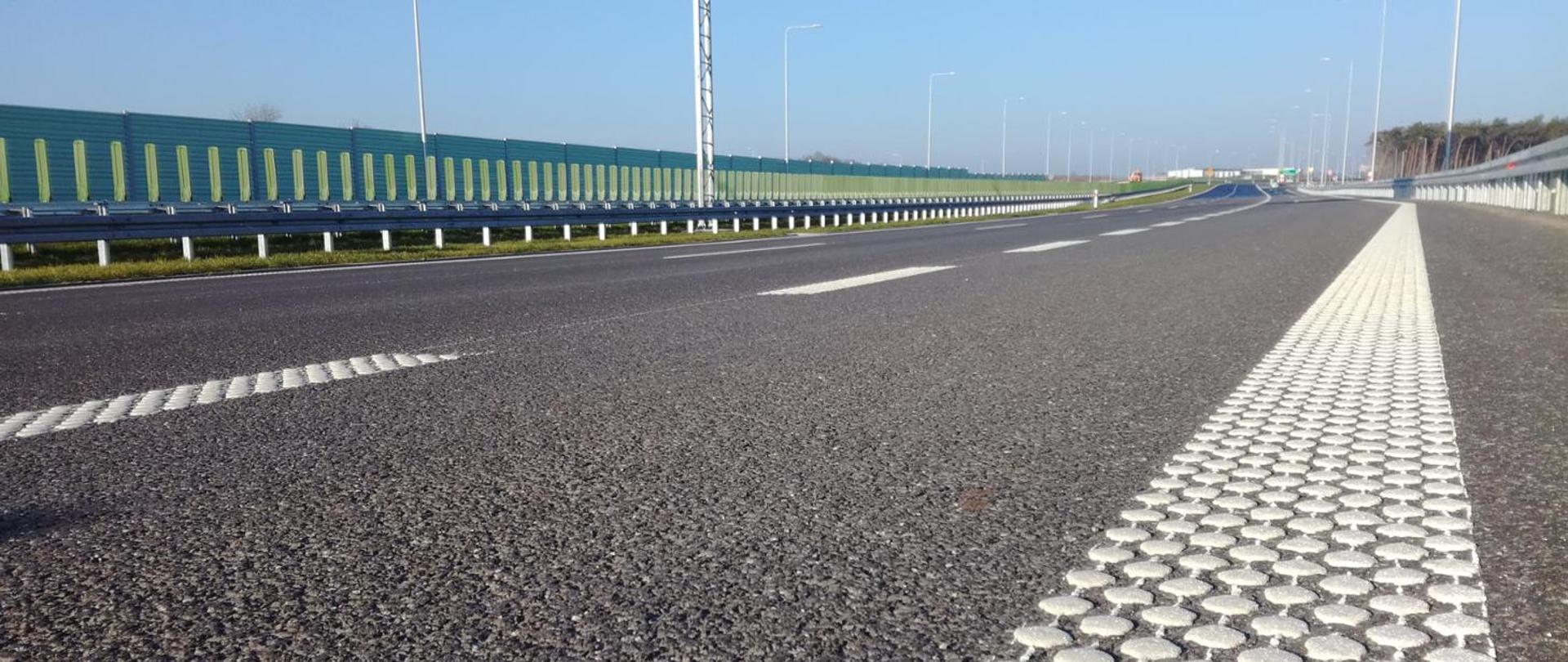 Zbliżenie na nawierzchnię drogi ekspresowej z widocznym pasem oznakowania krawędziowego i elementami odblaskowymi; w tle bariera energochłonna oraz ekrany akustyczne.