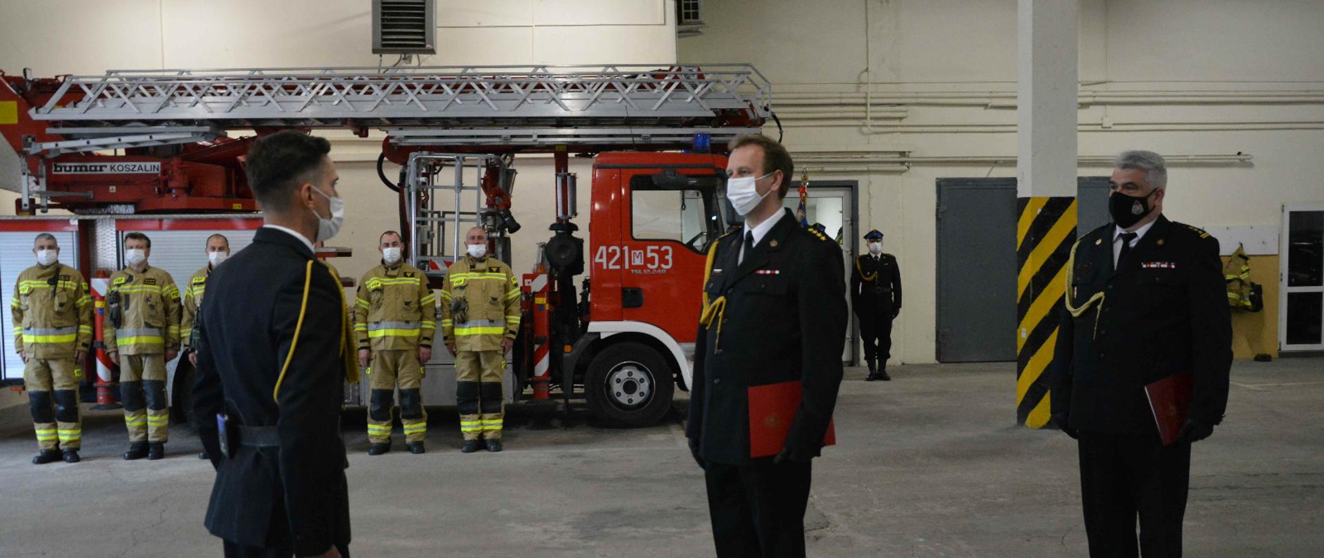 strażak składa meldunek przełożonemu. w tle strażacy w umundurowaniu bojowym i drabina pożarnicza. 