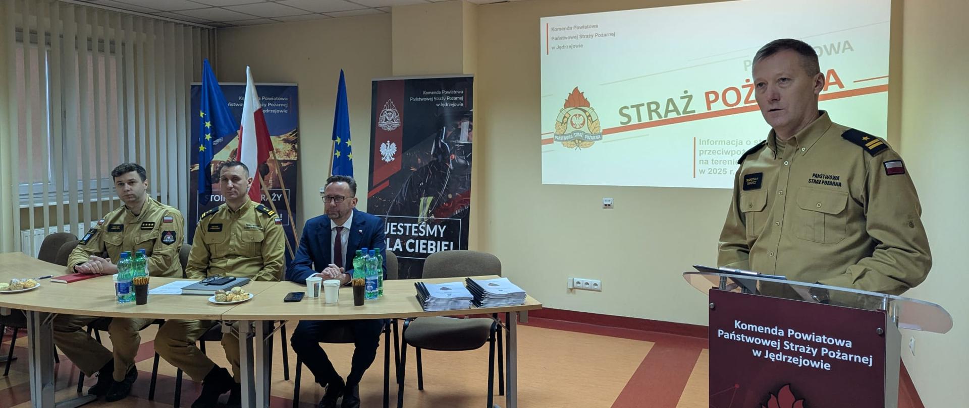 Przy stole prezydialnym widać od lewej zastępcę komendanta powiatowego PSP w Jędrzejowie st. kpt. Tomasza Kwietnia, zastępcę świętokrzyskiego komendanta wojewódzkiego PSP w Kielcach bryg. Michała Jankowskiego oraz starostę powiatu jędrzejowskiego Pana Pawła Farynę. Prezentację przedstawia komendant powiatowy PSP w Jędrzejowie mł. bryg. Sebastian Dudzicz.