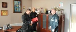 Nagrody Mazowieckiego Komendanta Wojewódzkiego PSP - w pokoju komendanta powiatowego stoi 3 strażaków w ubraniu służbowym, komendant czyta rozkaz