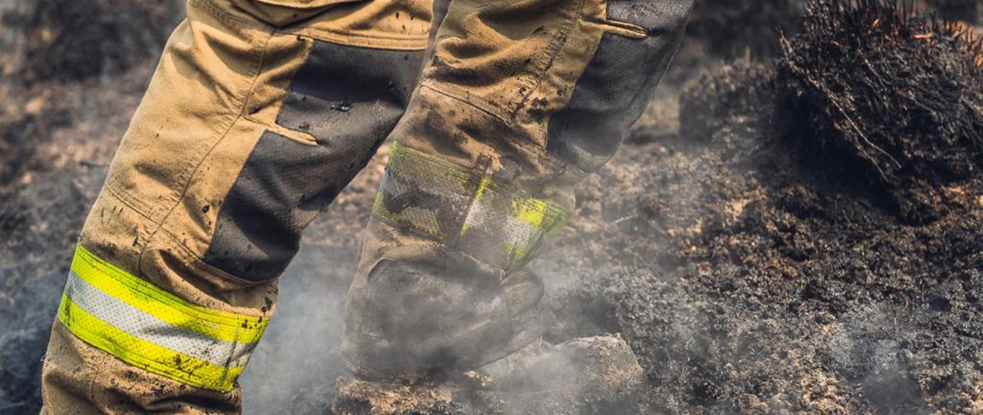 Na terenie wypalonym widoczne od kolan dwie nogi strażaka w spodniach specjalnych
