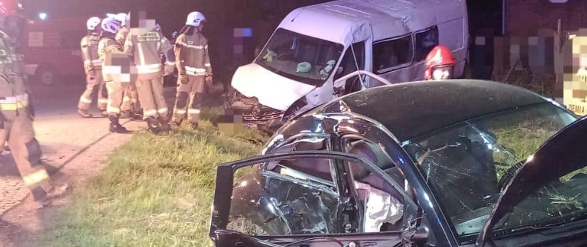 Na zdjęciu widać dwa rozbite samochody, biały bus oraz zielony osobowy. Oba pojazdy znajdują się na poboczu. Obok nich stoją strażacy w żółtych strojach i czerwonych lub białych hełmach. W tle widać budynek i czerwony samochód strażacki. Jest ciemno, ale miejsce akcji jest oświetlone.