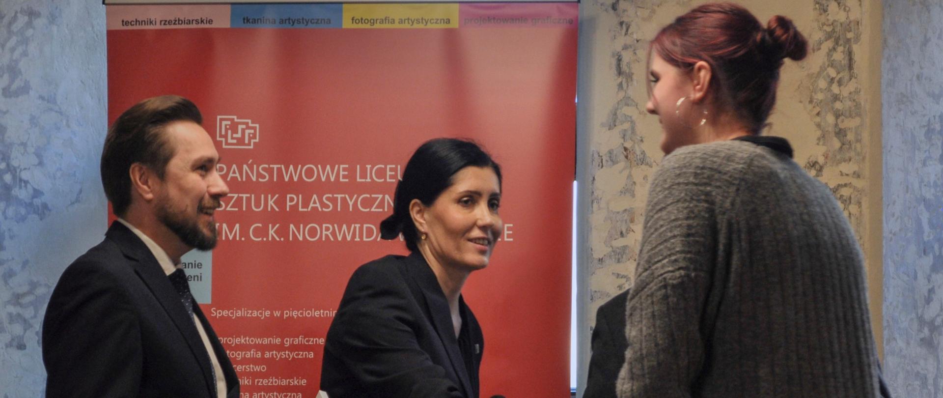 Po lewej stronie mężczyzna i kobieta, oboje elegancko ubrani. Po prawej młoda osoba, ściska dłoń kobiety. Za nimi czerwony baner Państwowego Liceum Sztuk Plastycznych im. C. K. Norwida w Lublinie