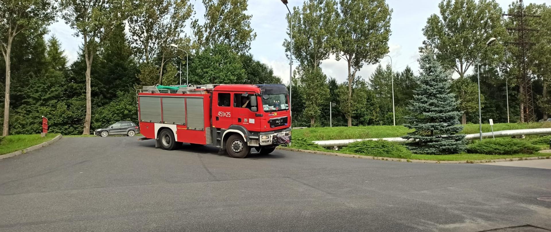 Pojazd koloru czerwonego. Na drzwiach widoczny kryptonim 451 - 25. Samochód ciężki ratowniczo gaśniczy wjeżdża na teren jednostki w tle drzewa 
