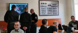 Przedstawiciele Państwowej Straży Pożarnej stoją przed ekranem telewizora patrząc na wyświetlaną mapę obok za stołami siedzą inni uczestnicy ćwiczeń.