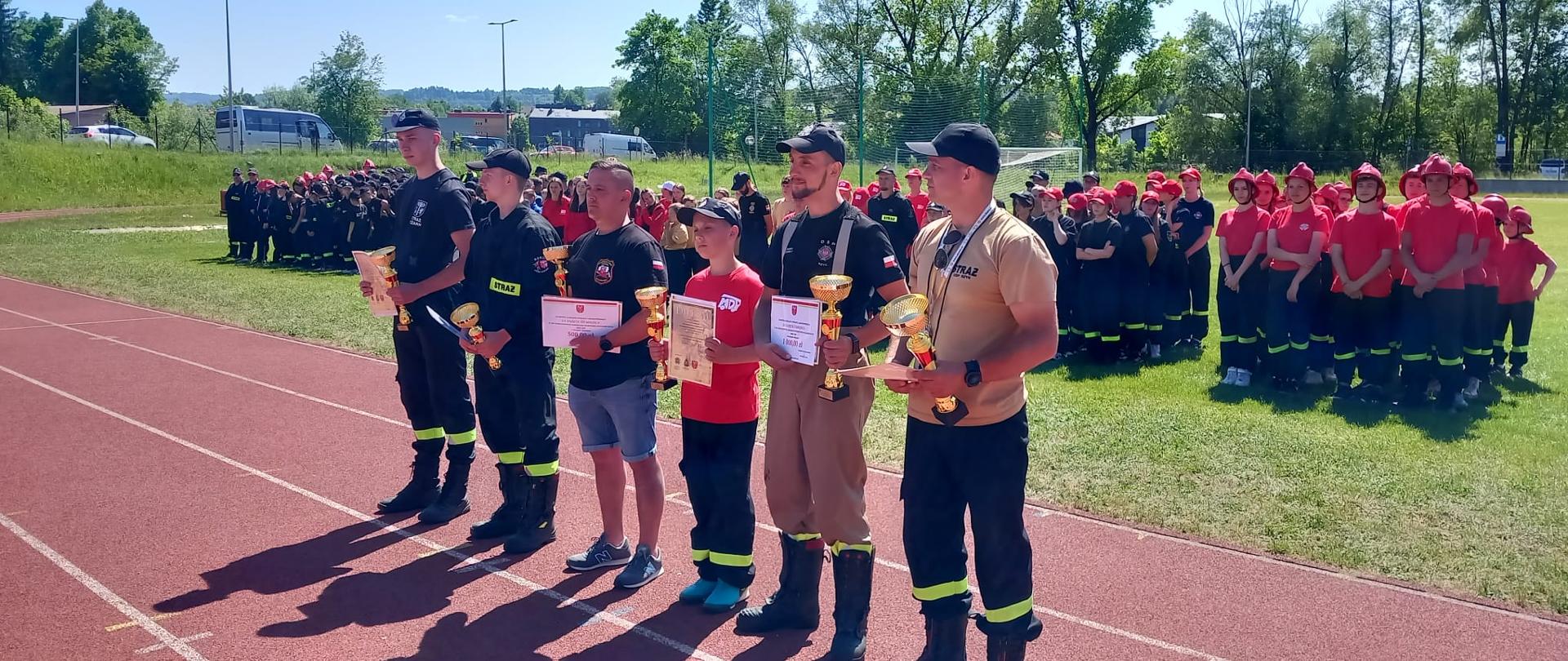 Zdjęcie przedstawia druhów OSP trzymających nagrody dla swoich jednostek OSP które stoją w tle. 
