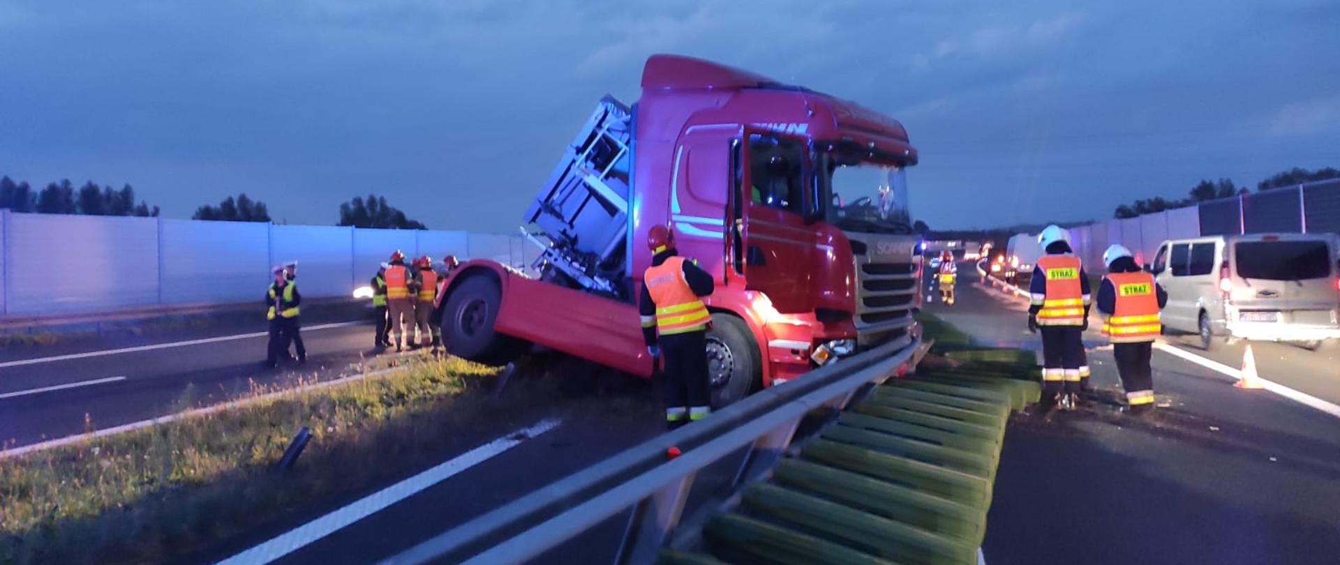 Na zdjęciu widać strażaków i funkcjonariuszy Policji, którzy prowadzą swoje czynności podczas usuwania skutków wypadku drogowego na Autostradzie A-4 Widoczny jest ciągnik siodłowy stojący w poprzek drogi pojazd koloru czerwonego, który znajduje się na pasie zieleni oddzielającym autostrady. 