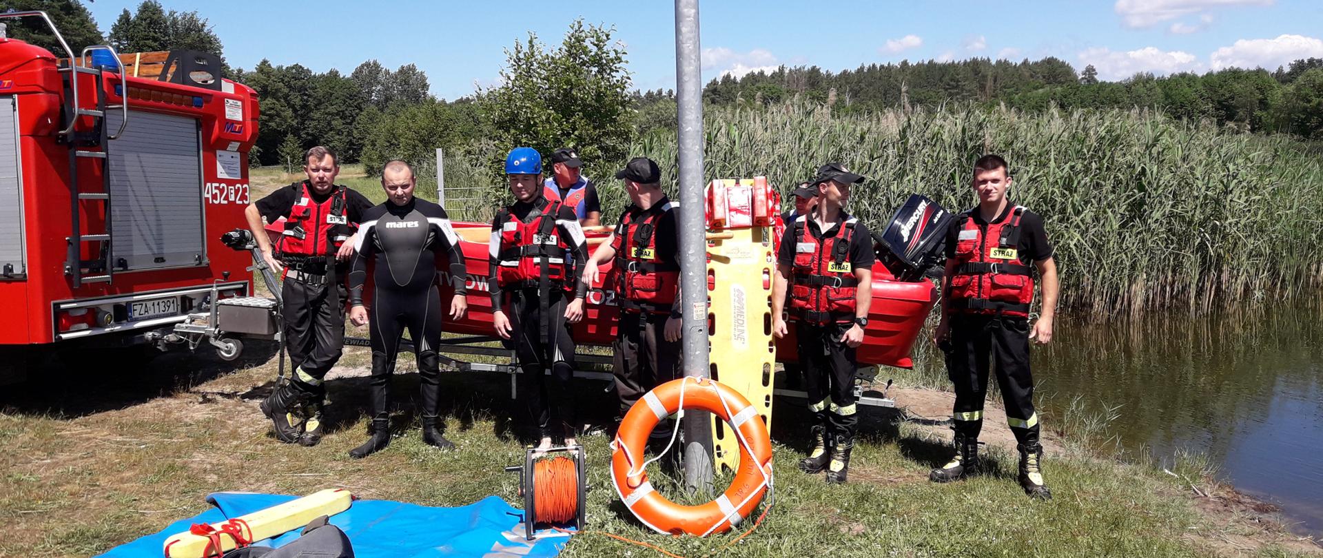 Zdjęcie wykonane nad zbiornikiem wodnym. W tle od lewej strony widoczny jest samochód specjalny obok podpięta łódź. Wokół łodzi stoi 8 strażaków ubranych w sprzęt do ratownictwa wodnego. Przed strażakami leży mata sprzętowa ze sprzętem do ratownictwa wodnego.