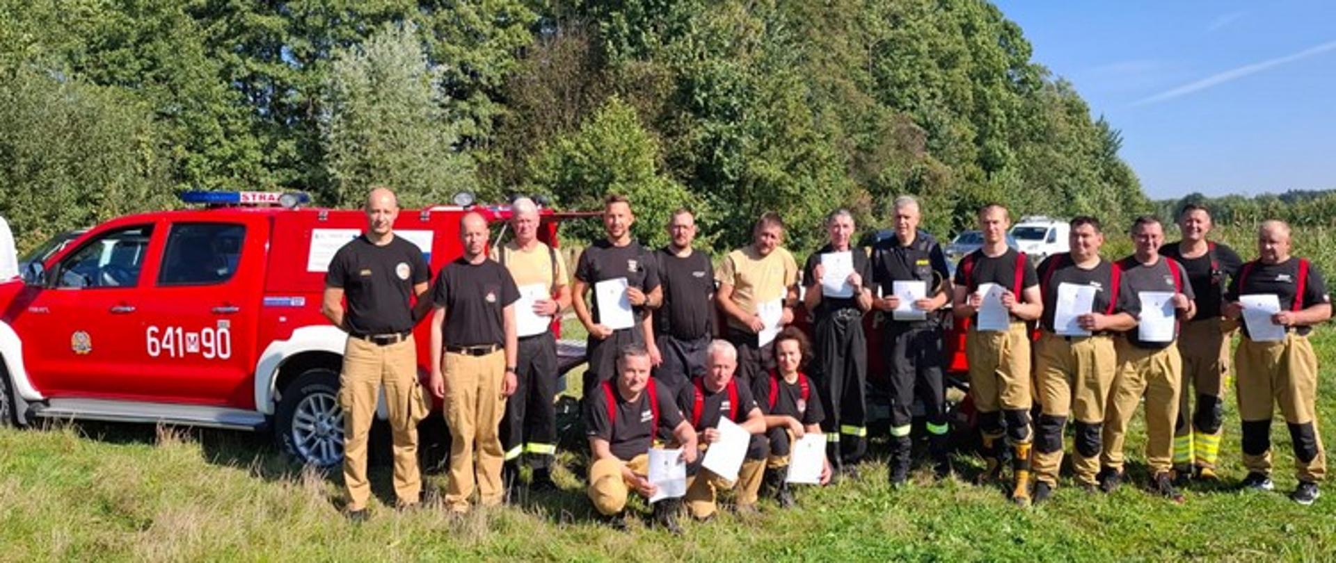 Zdjęcie przedstawia uczestników zakończonego kursu z ratownictwa wodnego. Osoby na zdjęciu ubrane w stroje koszarowe koloru czarnego i piaskowego. Trzymają w rękach zaświadczenia o ukończeniu kursu. Po lewej stronie widzimy samochód strażacki o kryptonimie 641m90. Zdjęcie wykonane na zielonej trawie w tle widoczny las liściasty. Pogoda na zdjęciu słoneczna, bez zachmurzeń.