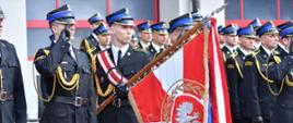 Na zdjęciu widoczny jest poczet sztandarowy Państwowej Straży Pożarnej podczas uroczystego apelu. Strażacy ubrani są w mundury galowe i stoją w szyku, a jeden z funkcjonariuszy trzyma sztandar jednostki ozdobiony barwami narodowymi oraz godłem państwowym. Pozostali strażacy oddają honor salutując podczas ceremonii. W tle znajdują się kolejni funkcjonariusze ustawieni przed budynkiem straży pożarnej.