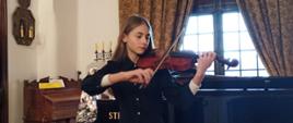Zdjęcie kolorowe. Jasnowłosa dziewczyna w czarnej sukience gra na skrzypcach. W tle okno ze szprosami częściowo przysłonięte ozdobnie upiętymi zasłonami, fortepian i zabytkowa fisharmonia.