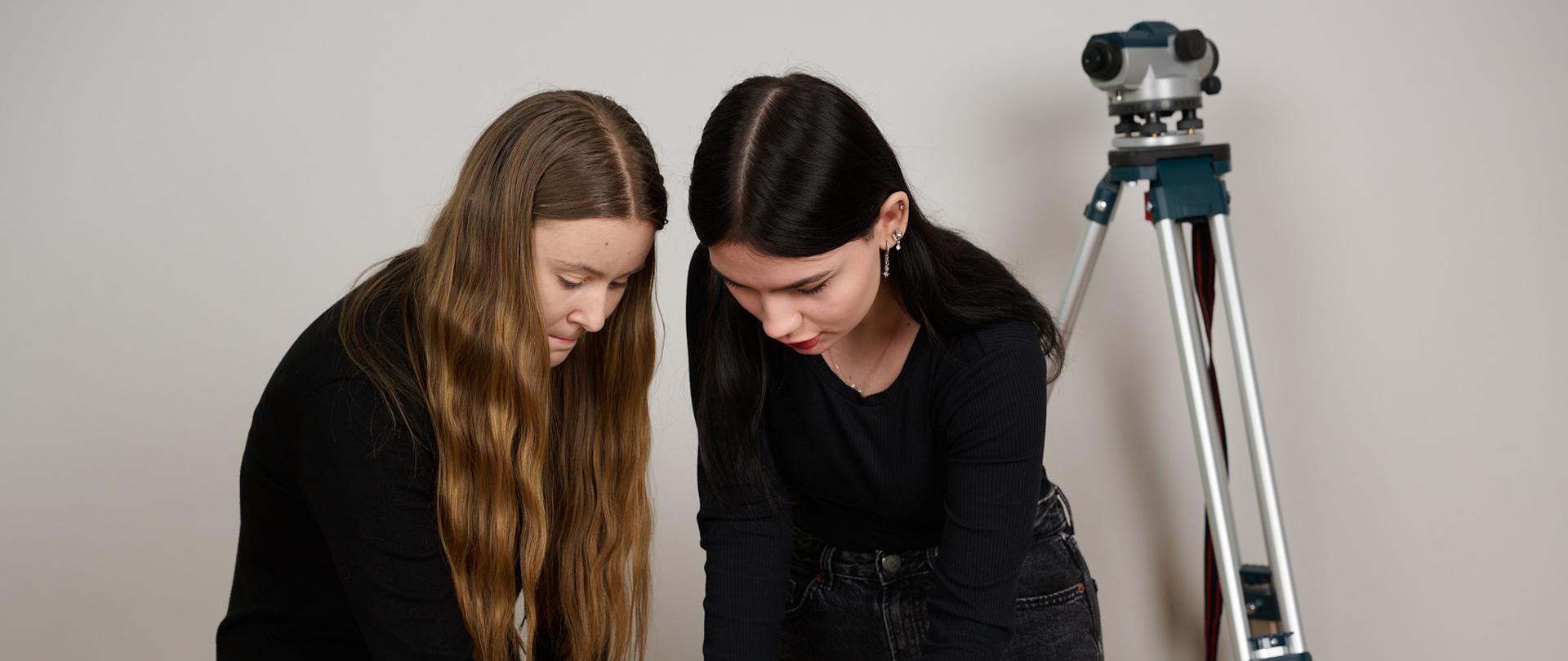 Dwie dziewczyny zaznaczają pisakami kartki z wydrukami zawierającymi wizualizacje projektową. Kartki leża na stoliku, w tyle rozłożony jest niwelator na statywie 