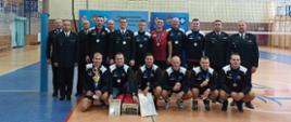 Na sali sportowej, na boisku do siatkówki drużyna sportowa pozuje do zdjęcia. Wśród pozujących są również strażacy w mundurach. 