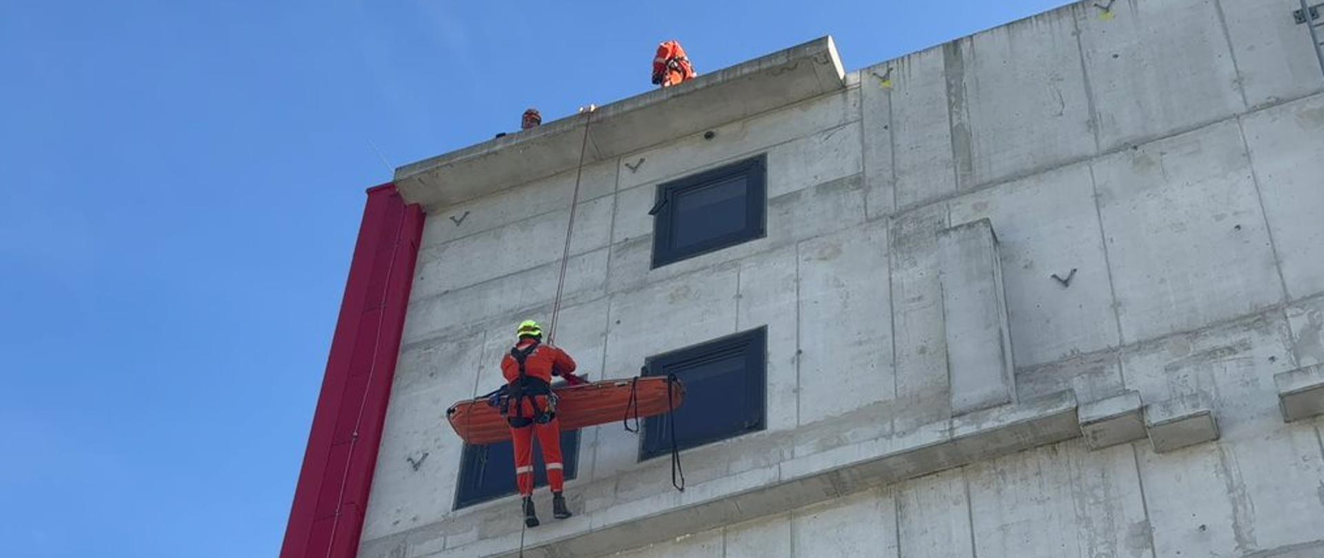 Strażacy w pomarańczowych kombinezonach. Jeden wisi na linach i trzyma pomarańczowe nosze. Dwóch stoi na dachu budynku