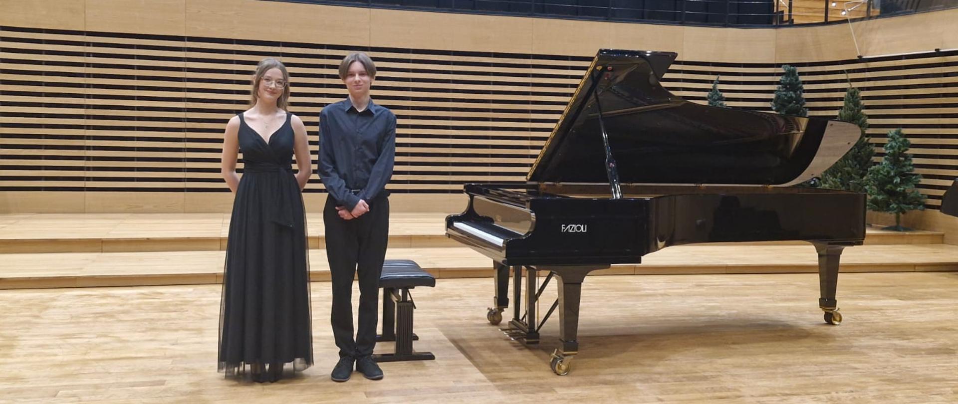 Chłopak i dziewczyna stoją na scenie przy fortepianie.