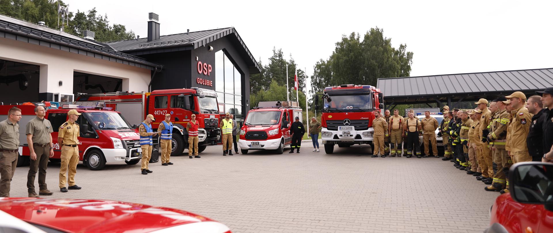 Zdjęcie przedstawia strażaków ustawionych na zbiórce, widać również ustawione wozy strażackie. W tle remiza OSP Gołubie
