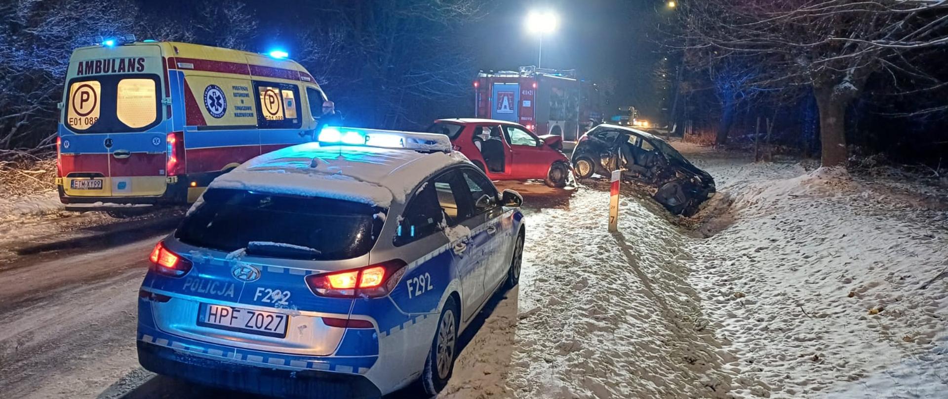 Zderzenie dwóch samochodów osobowych, droga i pobocza pokryte śniegiem, jeden czerwony samochód osobowy z uszkodzonym przodem stoi na jezdni, drugi pojazd osobowy z uszkodzonym przodem i prawym bokiem przodem pojazdu znajduje się przodem w przydrożnym rowie, na miejscu służby prowadzą działania, przed uszkodzonymi pojazdami stoi radiowóz policji, obok karetka pogotowia, za pojazdami stoi samochód strażacki z wysuniętym masztem, który oświetla teren na którym trwają działania ratownicze, ruch na drodze całkowicie zablokowany.