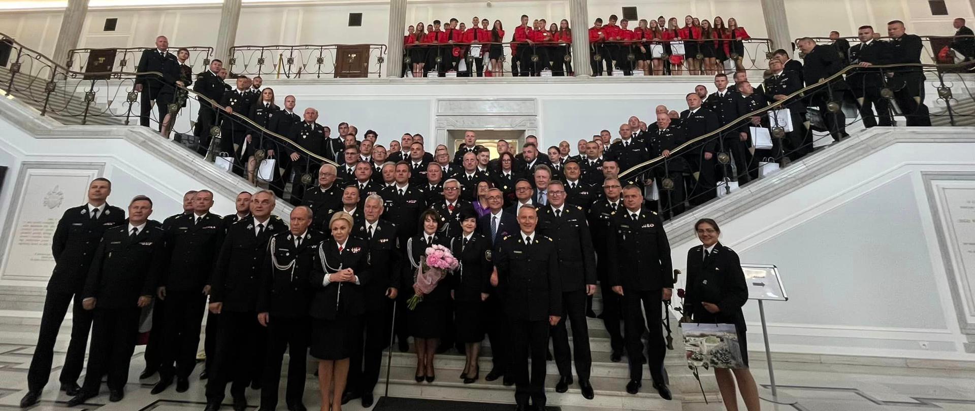 Zbiorowe zdjęcie uczestników konferencji - około 200 osób ustawionych na kształt krzyża, wzdłuż biegów schodów w gmachu Parlamentu. Na galerii młodzież w czerwonych mundurach.