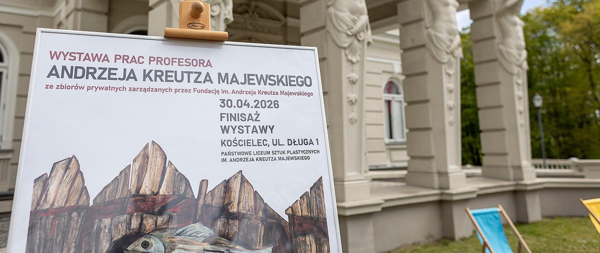 Bliskie ujęcie tablicy informacyjnej na drewnianej sztaludze, ustawionej przed zabytkowym pałacem. Napis na plakacie głosi: „Wystawa prac profesora Andrzeja Kreutza Majewskiego” z datą finisażu 30.04.2026. W tle widać fragment jasnej elewacji budynku ozdobionej rzeźbami kariatyd (postaci kobiecych podtrzymujących gzyms).